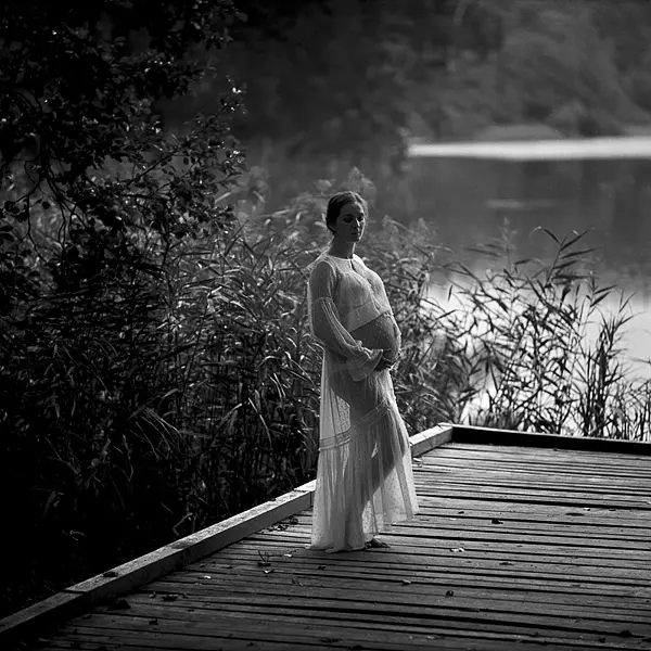 Stojąca na drewnianym pomoście młoda kobieta w ciąży na tle trzcin i jeziora