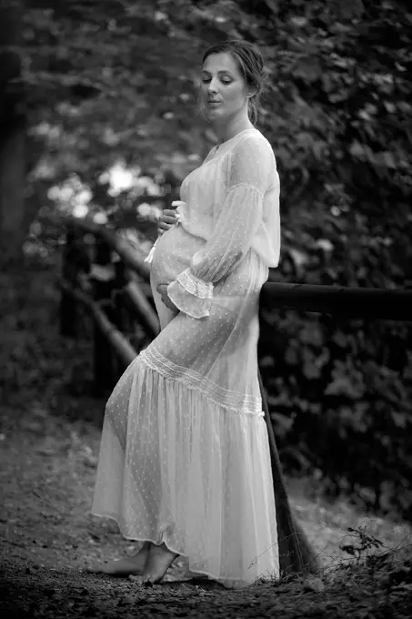 Młoda ciężarna kobieta w białej sukni oparta o drewnianą barierkę nad jeziorem