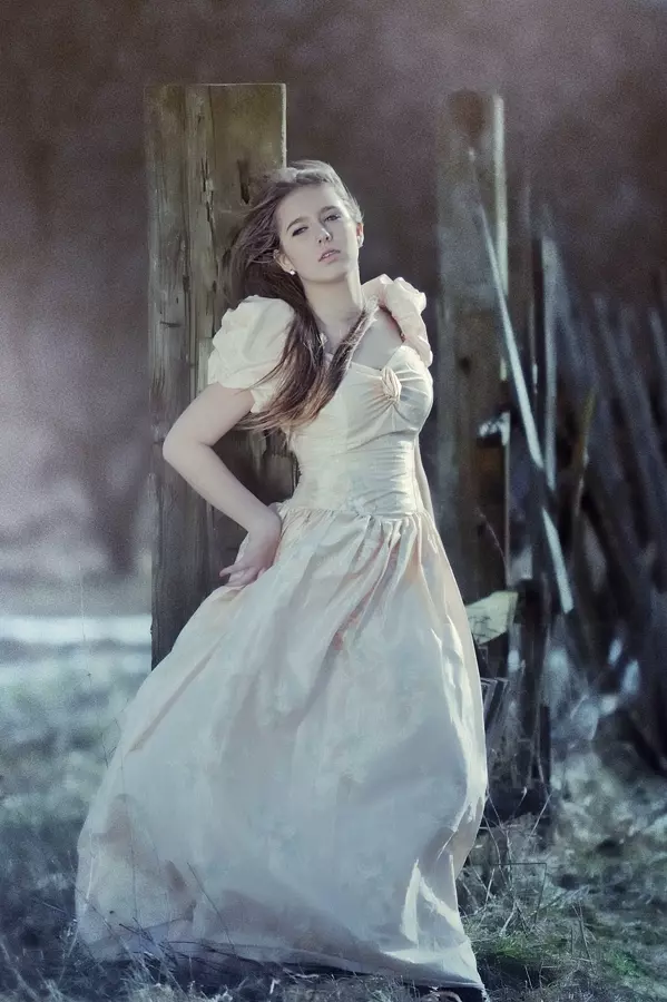 Modelka z rozwianymi na wietrze włosami pozująca przy starym płocie w łososiowej balowej sukni