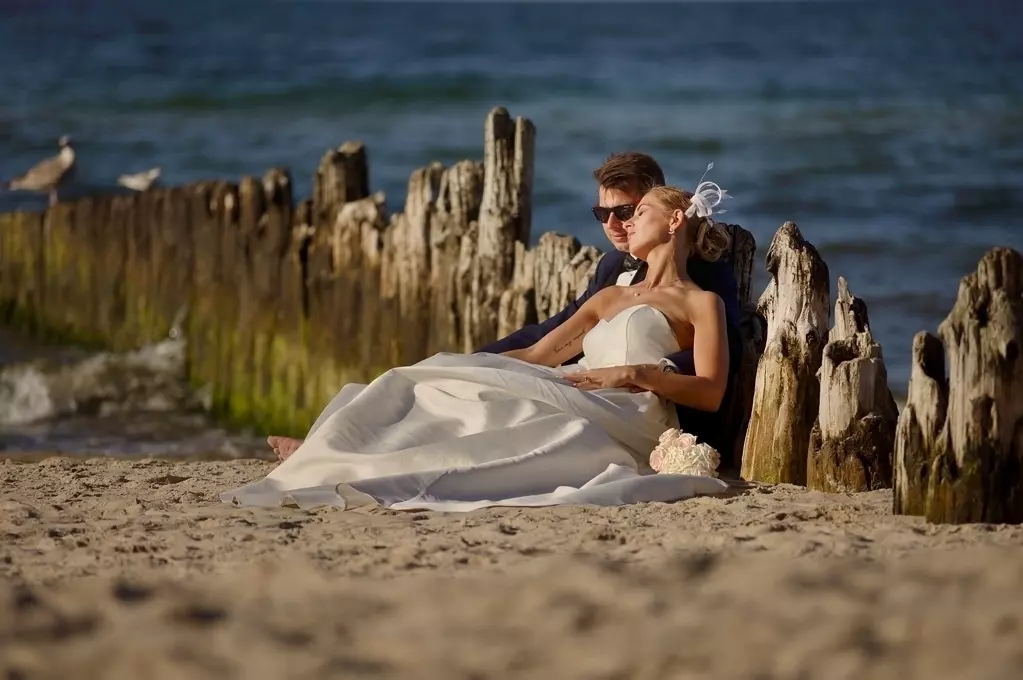 leżący na morskim piasku nowożeńcy pośród drewnianych falochronów